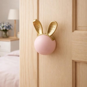 May include: A decorative door knob in the shape of a bunny. The knob has a pink sphere with gold-colored bunny ears. The knob is mounted on a light-colored wooden door. The background includes a bed and a lamp.