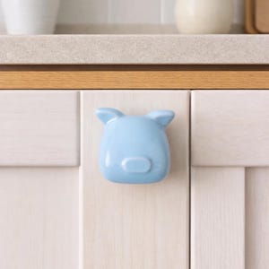以下が含まれることがあります： 水色の豚の形をしたキャビネットノブ。ノブは明るい色の木製キャビネットドアに取り付けられています。ノブには2つの小さな耳と丸みを帯びた鼻があります。背景には、カウンタートップ、白いタイル、花瓶の花が含まれています。