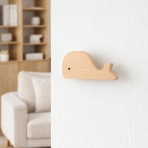 Może przedstawiać: Drewniany haczyk ścienny w kształcie wieloryba. Haczyk wykonany jest z jasnego drewna i ma prosty, minimalistyczny design. Jest zamontowany na białej ścianie i nadaje się do pokoju dziecięcego.