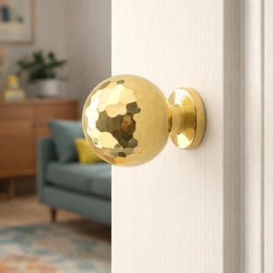Könnte beinhalten: Ein goldfarbener Türknauf mit gehammerter Textur und runder Form. Der Knauf ist an einer weißen Tür montiert, mit einem Blick auf ein Wohnzimmer im Hintergrund.