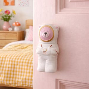 Peut inclure: Bouton de porte en forme d'ours blanc avec un visage rose et des accents dorés. L'ours porte une combinaison spatiale et est monté sur une porte rose. L'arrière-plan comprend un lit avec une couette à carreaux jaunes et blancs.