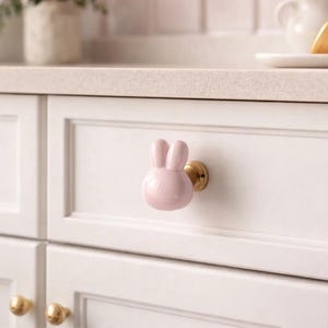 May include: A pink ceramic bunny-shaped cabinet knob with a gold base is attached to a white cabinet door. The knob is a decorative accent for kitchen or bathroom furniture. Other gold knobs are visible below.