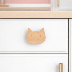 May include: A wooden cat-shaped drawer pull is mounted on a white cabinet door. The pull has two small holes for eyes and a light brown finish. A gold-colored handle is visible below. The cabinet is part of a kitchen.