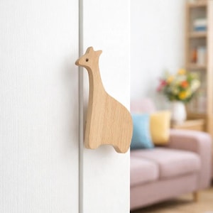 Peut inclure: Poignée de porte en forme de girafe en bois clair, fixée sur une porte blanche. Le design est simple et minimaliste. La tête et le cou de la girafe sont allongés, avec un petit trou pour l'œil.