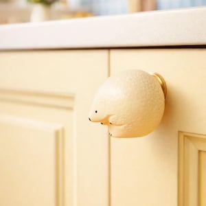 May include: A cream-colored hedgehog-shaped cabinet knob with a textured surface and black eyes. The knob is mounted on a light yellow cabinet door with a gold-colored base. The knob adds a whimsical touch to kitchen or bathroom cabinetry.
