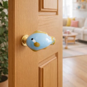 Könnte beinhalten: Ein blau-gelber Türknauf in Fischform aus Keramik mit Messingfuß ist an einer hellbraunen Holztür befestigt. Die Tür ist teilweise geöffnet und gibt den Blick auf ein Wohnzimmer mit Sofa und Pflanze frei.