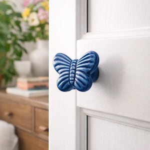 Puede incluir: Pomo de puerta de cerámica azul con forma de mariposa. El pomo está montado en una puerta blanca. El diseño de la mariposa tiene patrones de alas detallados y una superficie texturizada. El pomo añade un toque decorativo a cualquier puerta.