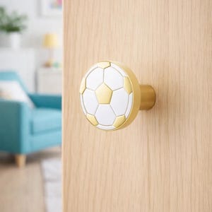 May include: A decorative door knob with a soccer ball design. The knob is white and gold with a gold base, mounted on a light wood door. The soccer ball pattern is made up of white and gold pentagons and hexagons.