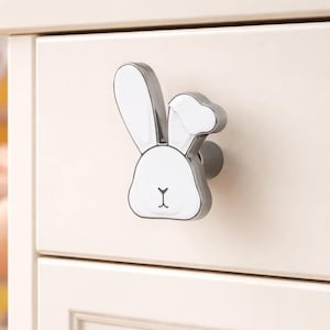 Puede incluir: Tirador de cajón con forma de conejo blanco y plateado. La cara del conejo es blanca con un contorno negro y una pequeña nariz negra en forma de 'Y'. Montado en un cajón de color claro, sugiere un tema infantil o de guardería.