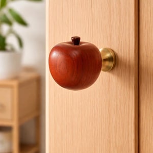 Op de afbeelding: Een houten deurknop in de vorm van een appel met een donkerrode afwerking en een kleine bruine steel. De knop is gemonteerd op een lichtgekleurde houten deur met een messing basis. Decoratieve hardware.