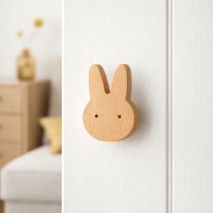 Puede incluir: Un gancho de pared de madera con forma de conejito. El gancho es de madera clara y presenta un diseño sencillo con dos pequeños agujeros para los ojos. Está montado en una puerta blanca, añadiendo un toque de fantasía a cualquier habitación.