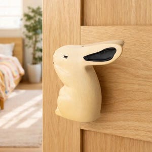 May include: A light beige bunny-shaped door stop made of wood. The bunny has black accents on its ears and closed eyes. The door stop is mounted on a light-colored wooden door.