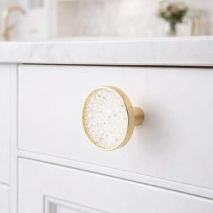 May include: A gold cabinet knob with a round, white, crystal-like center. The knob is attached to a white cabinet door. The background is a blurred white kitchen setting with a marble countertop.