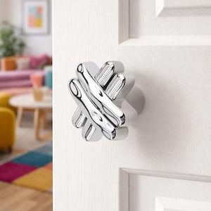 以下が含まれることがあります： ユニークな十字型のデザインの、磨かれたクロム製のドアノブ。白いドアに取り付けられており、背景にはカラフルなインテリアが垣間見えます。ノブの直径は約10cmです。