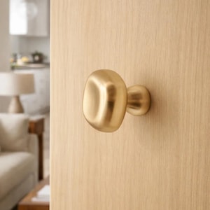 以下が含まれることがあります： 丸みを帯びた有機的な形状の金色のドアノブが、明るい色の木製のドアに取り付けられています。ノブは滑らかで磨かれた仕上げで、小さな円形のベースが付いています。ドアは部分的に開いており、生活空間を垣間見ることができます。