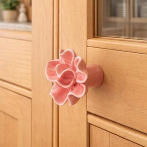 May include: A pink ceramic flower-shaped cabinet knob. The knob is mounted on a light-coloured wooden cabinet door. The flower has multiple ruffled petals and a rounded base. The cabinet door is made of light-coloured wood.