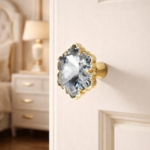 May include: A decorative door knob with a clear, faceted crystal centre and a gold-coloured metal base. The knob is flower-shaped and is mounted on a white door. The background includes a bedroom setting.
