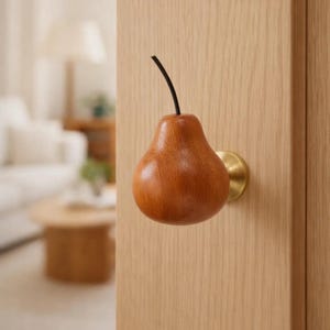 Pode incluir: Um puxador de porta em forma de pêra de madeira, com base em latão e haste preta. A pêra é de cor castanha quente e está montada numa porta de madeira clara. O puxador adiciona um toque de fantasia à porta.