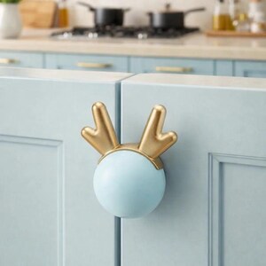 May include: A light blue cabinet knob with a gold antler design. The knob is round with a gold antler design on top. The knob is mounted on a light blue cabinet door. Kitchen decor.