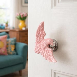 May include: A pink ceramic door knob in the shape of a bird's wing is mounted on a white door. The wing has detailed feather textures and is attached to a silver-colored metal base.