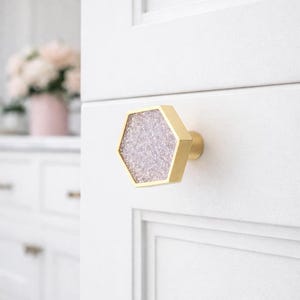 May include: A gold-toned, hexagonal cabinet knob with a glittery, light purple center. The knob is mounted on a white cabinet door. The design is modern and decorative, suitable for kitchen or bathroom cabinetry.