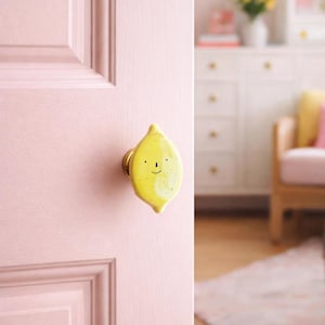 Könnte beinhalten: Ein gelber, zitronenförmiger Türknauf mit einem lächelnden Gesicht ist an einer rosa Tür angebracht. Der Knauf hat einen goldfarbenen Sockel. Die Tür ist Teil eines Raumes mit einer weißen Kommode und einem Holzstuhl.