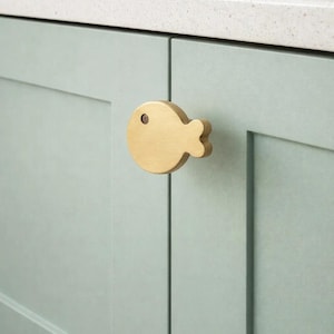 Könnte beinhalten: Ein goldfarbener, fischförmiger Schrankknauf ist an einer hellgrünen Schranktür befestigt. Der Knauf hat einen kreisförmigen Körper mit einem kleinen Augendetail und einer Schwanzflosse. Die Schranktür ist Teil eines Küchenschranksatzes.