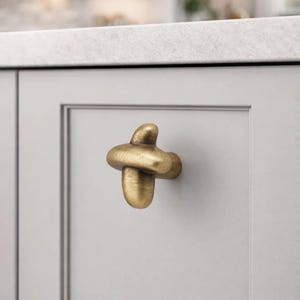 May include: A brass-colored cabinet knob with a unique, knotted design. The hardware is mounted on a light gray cabinet door, offering a modern touch to kitchen or bathroom decor. The knob's finish has a slightly aged appearance.