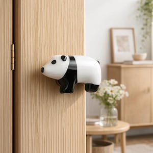 May include: A panda-shaped door knob in white and black is fixed to a light-coloured wooden door. The door knob features black eyes, ears, and feet, with a black stripe across its back. The door is attached to a gold-coloured hinge.