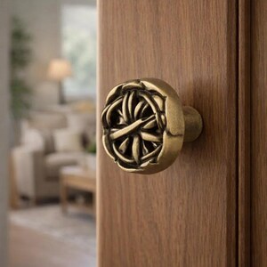May include: Close-up of a bronze-colored door knob with an intricate, woven design. The knob is mounted on a wooden door, with a blurred interior visible in the background. The door knob adds a decorative touch to the door.