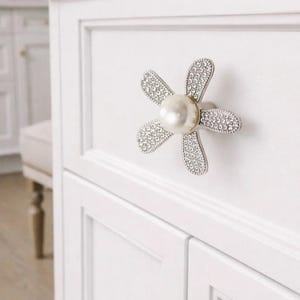 Puede incluir: Un tirador de armario con forma de flor, con un centro de perla y detalles de cristal. El tirador plateado está montado en una puerta de armario blanca. Diseño elegante y decorativo.