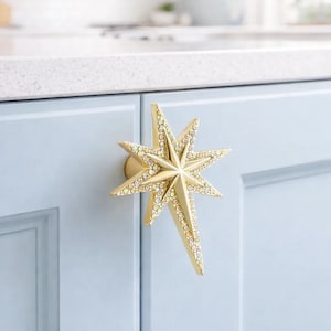 Pode incluir: Um puxador de armário dourado em forma de estrela com detalhes em cristal. O puxador está montado numa porta de armário azul claro. O design em estrela adiciona um toque decorativo.