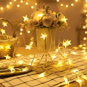 May include: A warm, inviting scene featuring star-shaped fairy lights draped around a floral arrangement in a white vase. The lights are strung across a checkered tablecloth, creating a cozy ambiance. A basket is visible in the background.