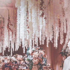 May include: A wedding or event venue decorated with cascading white and pink floral garlands suspended from the ceiling. The garlands are interspersed with small pink flowers. Floral arrangements in shades of pink and peach are visible on tables.