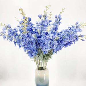 May include: A bunch of blue delphinium flowers in a white and blue vase. The flowers have multiple layers of petals and are a vibrant shade of blue. The vase is cylindrical and has a gradient colour scheme.