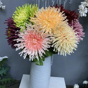 Puede incluir: Un ramo de crisantemos artificiales coloridos en un jarrón blanco. Las flores son en tonos de rosa, melocotón, amarillo, verde y burdeos. El jarrón tiene un diseño texturizado y acanalado. El arreglo está sobre una superficie blanca.