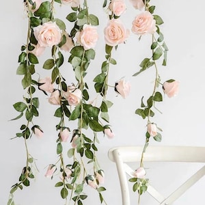 Op de afbeelding: Kunstmatige rozenranken met lichtroze bloesems en groene bladeren hangen naar beneden. De rozen zijn in verschillende stadia van bloei, van gesloten knoppen tot volledig geopende bloemen. De ranken hangen tegen een witte achtergrond, met een witte stoel in de rechter benedenhoek.