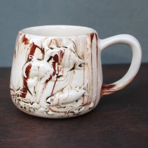 May include: A ceramic mug with a white base and a brown, marbled design. The mug features a raised relief of a forest scene with animals. The handle and interior are white, creating a contrast with the exterior.