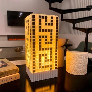 May include: A modern, illuminated lamp shaped like a skyscraper, featuring a grid pattern of black squares on a yellow background. The lamp sits on a white base and is placed on a black surface. Books are stacked nearby.