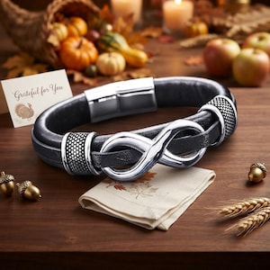 Puede incluir: Una pulsera de cuero negro con un símbolo de infinito en tono plateado pulido y dos detalles metálicos texturizados en tono plateado. La pulsera incluye un cierre magnético rectangular plateado y se muestra con una tarjeta que dice Grateful for You.
