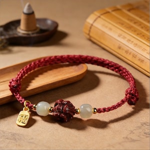 May include: A red braided bracelet with a gold charm and decorative beads. The charm has Chinese characters. The bracelet features a carved dark red bead and two light green beads. The bracelet is displayed on a wooden surface.