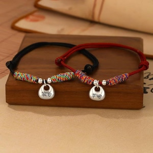 Può includere: Due braccialetti con un ciondolo d'argento. Uno è nero, l'altro rosso. I ciondoli hanno caratteri cinesi. I braccialetti hanno sezioni intrecciate colorate. Sono su una superficie di legno.