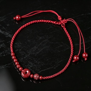 May include: A red braided cord bracelet featuring a central circular donut-shaped bead, a single textured spherical bead, and several smaller smooth round beads. The bracelet includes adjustable sliding knots with two dangling ends finished with beads.