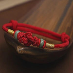 May include: Red braided bracelet with a central knot design, gold accents, and two jade-colored beads. The bracelet is made of a thick, woven red cord. The gold accents are wrapped around the cord. The bracelet is displayed on a wooden surface.