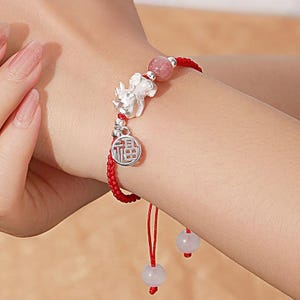 Peut inclure: Bracelet tressé rouge avec des breloques argentées. Il présente une figurine Pixiu, un charm rond avec des caractères chinois et des perles roses et blanches. Le bracelet est noué avec un nœud coulissant pour un ajustement.