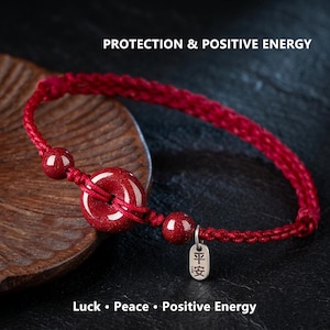 Può includere: Braccialetto rosso intrecciato con una perla centrale circolare e due perline più piccole. Un piccolo ciondolo d'argento con caratteri cinesi è appeso. Il testo "PROTECTION & POSITIVE ENERGY" e "Luck • Peace • Positive Energy" sono visibili.