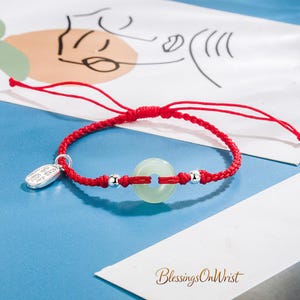 May include: Red braided bracelet featuring a pale green jade donut pendant, silver beads, and a small silver charm. The bracelet is displayed on a blue surface with a white paper with abstract art. The bracelet is adjustable.
