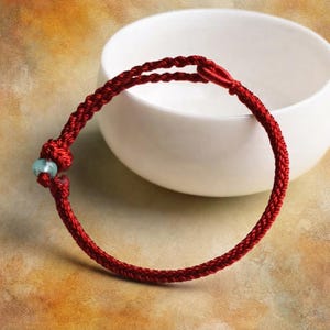 May include: A red braided cord bracelet featuring a single light blue bead. The bracelet is constructed from a continuous length of twisted red thread, secured with a knot near the bead, forming a circular band with a simple, functional design.
