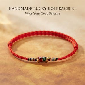 May include: A red braided cord bracelet featuring a central metal koi fish charm with red, green, and gold enamel details. The text HANDMADE LUCKY KOI BRACELET and Wear Your Good Fortune are printed above the bracelet.