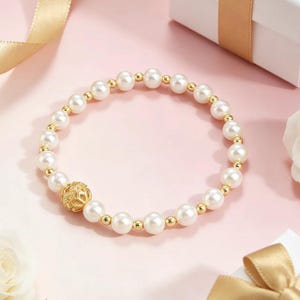 Op de afbeelding: Een parelarmband met gouden accenten en een decoratieve gouden kraal. De armband heeft ronde, witte parels en kleine gouden kralen. De decoratieve kraal heeft een open ontwerp. De armband wordt gepresenteerd op een roze ondergrond.